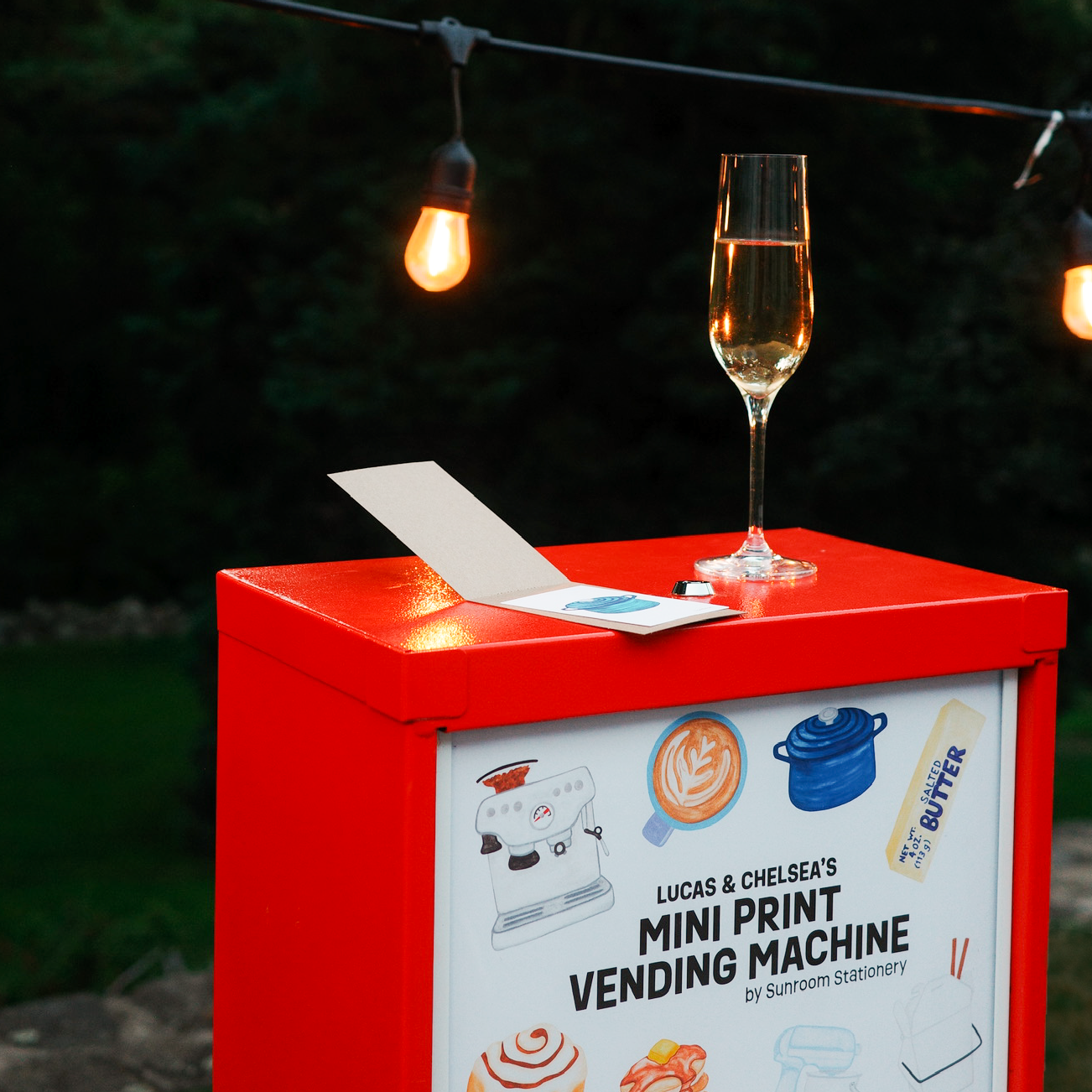 A red mini print vending machine with a white front panel stands outdoors at night under warm string lights. A glass of champagne rests on top next to a small card.